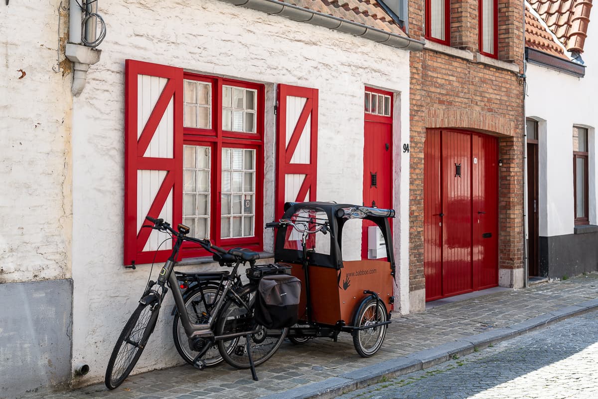 1-2 kleine Häuschen in Brügge, mit roten Fensterrahmen und rot-weißen Fensterläden, einer roten Tür und einem roten Tor in der Einfahrt. Davor ein Fahhrad und ein weiteres davor mit einem überdachten Fahrradanhänger. 