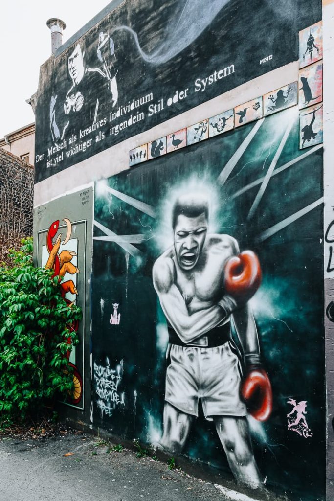 Graffiti im Unionviertel in Dortmund. Muhammad Ali in K.O.-Pose mit Boxhandschuhen und Shorts - er blickt nach unten auf einen nicht zu sehenden Gegner, der linke Arm am Körper ausgestreckt, die rechte angewinkelt über Bauch und Brust. Scheinwerfer sind auf ihn errichtet und er bekommt dadurch einen Heiligenschein.