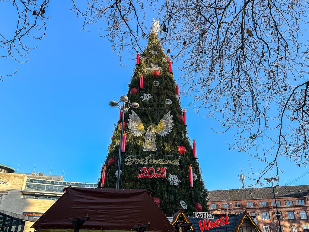 Auf einem Platz steht ein hoher Weihnachtsbaum, geschmückt mit roten Kerzen, roten und goldenen Ornamenten und einem 2021-Schild. Im Vordergrund steht eine kleine Hütte mit der Aufschrift „WEITERE Würst“. Kahle Äste rahmen die Szene vor einem klaren blauen Himmel ein, im Hintergrund sind Gebäude zu sehen.