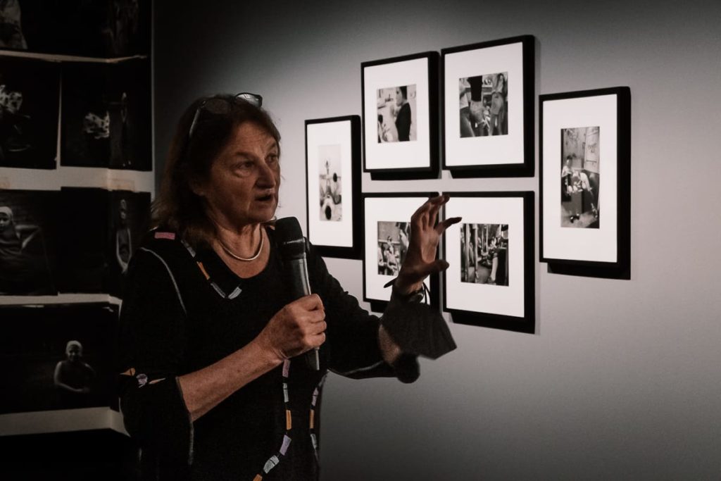 Eine Frau mit einem Mikrofon deutet auf eine Wand mit sechs gerahmten Schwarzweißfotos. Sie steht in einer schwach beleuchteten Galerie, die nur teilweise beleuchtet ist. An der Wand links von ihr sind weitere Fotos zu sehen.

Susan Meiselas vor Bildern aus ihrem Frühwerk im C/O Berlin.