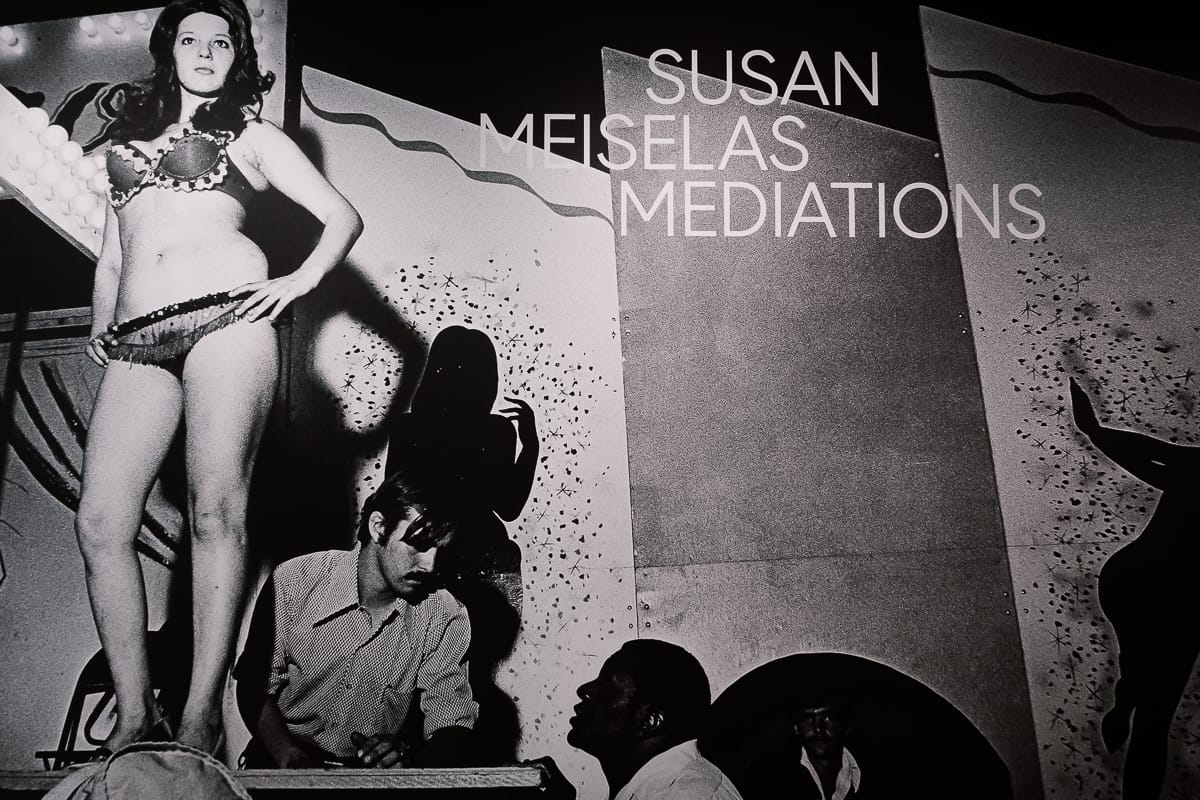 Schwarz-Weiß-Foto einer Frau im Bikini, die neben abstrakten Tafeln auf der Bühne steht, mit zwei Männern unter ihr. Über dem Bild steht in großem Schriftzug „SUSAN MEISELAS MEDIATIONS“. Die Szene scheint einer Aufführung oder einem Nachtclub zu entstammen. Susan Meiselas - Mediations, Ausstellungsmotiv mit Schriftzug darauf, C/O Berlin.