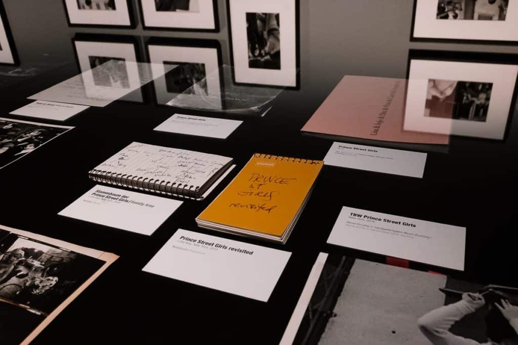 Vitrine mit verschiedenen Notizbüchern, Fotos und Bildunterschriften auf einer schwarzen Fläche. In der Mitte befindet sich ein gelber Notizblock mit dem Titel „Prince St. Girls revisited“. Im Hintergrund hängen gerahmte Schwarz-Weiß-Fotos an der Wand.

Notizhefte, Briefe, Kassetten zu Prince Street Girls und anderen Konzeptarbeiten von Susan Meiselas im C/O Berlin.
