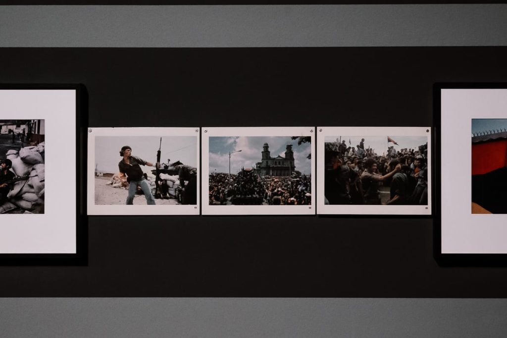An einer Galeriewand hängen vier historische Fotografien. Das linke Bild zeigt Sandsäcke, das zweite eine Person mit einem Gewehr, das dritte eine dichte Menschenmenge und das vierte eine Gruppe mit erhobenen Armen. Die einzelnen Fotos sind durch Ränder voneinander getrennt und stehen auf einem dunklen Hintergrund.