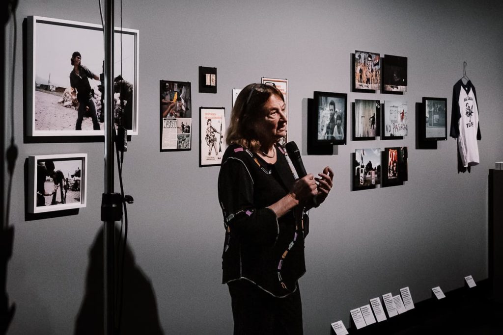 Eine Frau mit langen Haaren hält ein Mikrofon und spricht vor einer grauen Wand, an der gerahmte Fotos, Zeitungsausschnitte und ein T-Shirt hängen. Das Licht ist auf sie gerichtet, während im Hintergrund die Bilder und Schaukarten zu sehen sind.

Susan Meiselas im C/O Berlin vor ihrem Bild des Molotov Man.