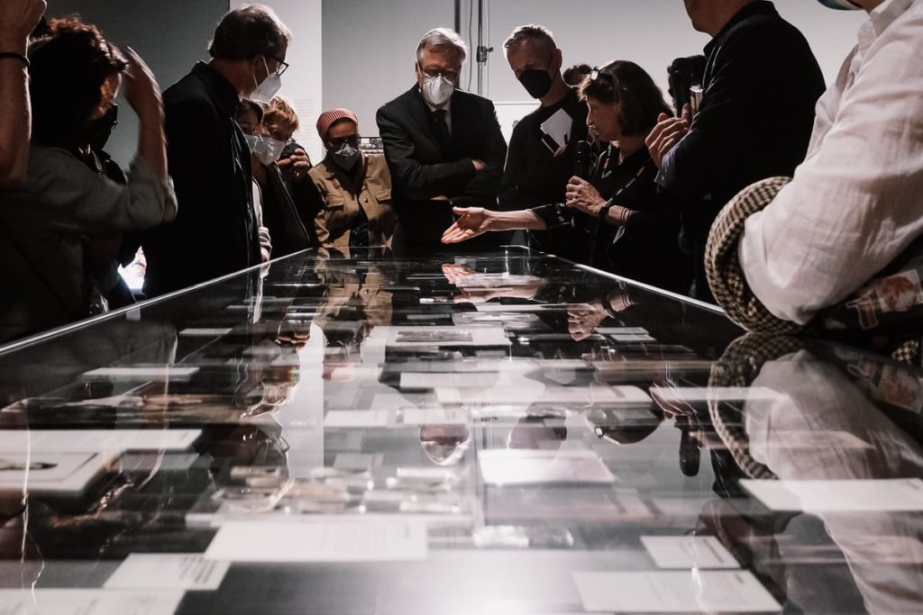 Eine Gruppe maskierter Menschen steht um eine Glasvitrine herum und betrachtet Dokumente und Fotos darin. Eine Person gestikuliert beim Sprechen und suggeriert damit eine Führung oder Diskussion in einem Museum oder einer Ausstellung. Die Beleuchtung ist auf die Vitrine gerichtet.

Susan Meiselas erklärt über einer Vitrine die Auseinandersetzung mit der kurdischen Diaspora.