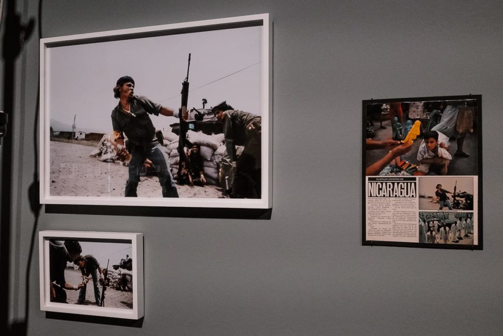 Gerahmte Bilder an einer grauen Wand zeigen bewaffnete Männer in einer militärischen Umgebung, Sandsäcke sind zu sehen. Daneben hängt auch eine Zeitschriftenseite mit Fotos, Text und der Schlagzeile „NICARAGUA“. Die Szene vermittelt Themen wie Konflikt und historische Dokumentation.