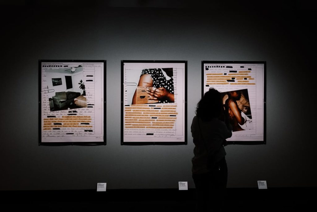 Eine Person mit lockigem Haar steht als Silhouette da und betrachtet drei gerahmte Kunstwerke an einer dunklen Galeriewand. Jedes Kunstwerk kombiniert Fotos und Dokumente mit großen, geschwärzten Textabschnitten, die Themen wie Privatsphäre oder Zensur verdeutlichen. Unter jedem Rahmen befinden sich kleine Schilder.

Bilder aus Archives of Abuse bzw. A Room of Their Own, Susan Meiselas.