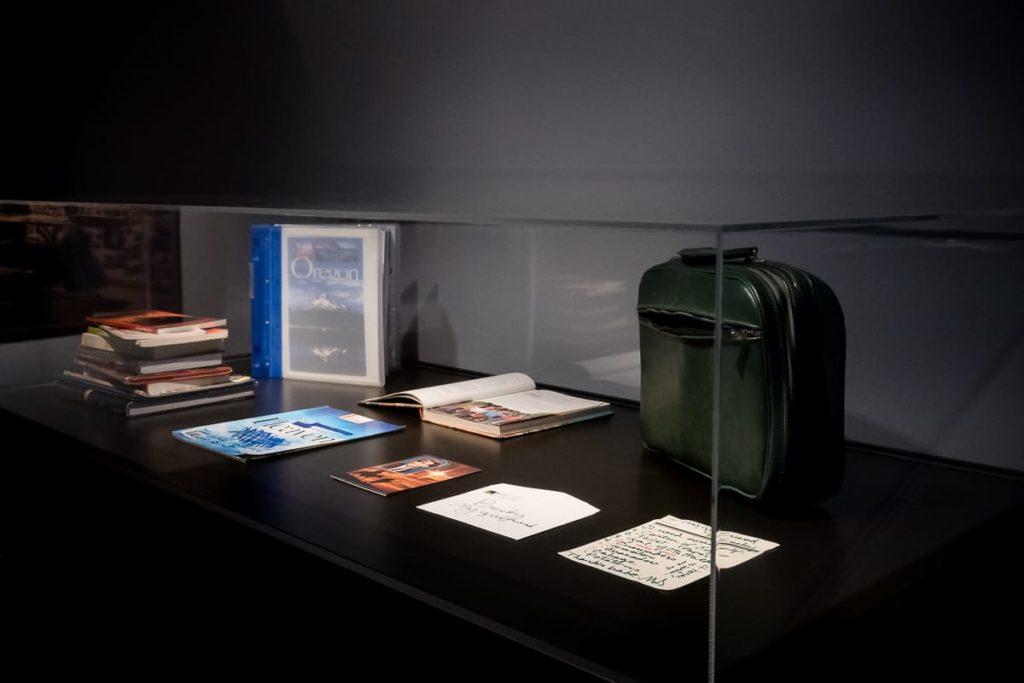 In einer Museumsvitrine liegen gestapelte Bücher, ein blauer Ordner, eine aufgeschlagene Zeitschrift, Postkarten, handschriftliche Notizen und ein kleiner dunkelgrüner Koffer, alles ordentlich auf einer schwarzen Fläche unter Glas angeordnet. Der Hintergrund ist schwach beleuchtet.

Überbleibsel einer Begegnung und Spurensuche, Michaels Koffer und Bücher. Bieke Depoorter, C/O Berlin.