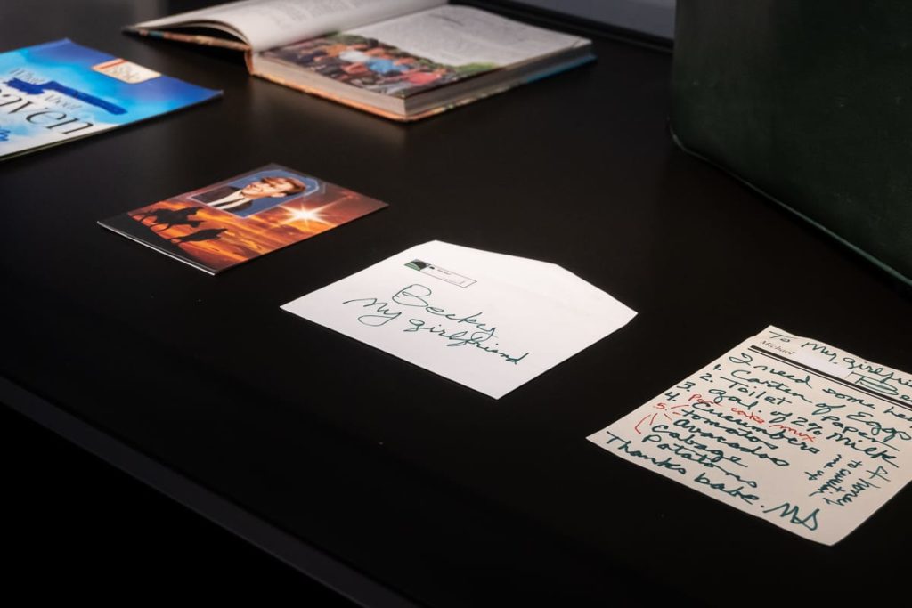 Eine Nahaufnahme eines schwarzen Tisches mit einem Foto, einem handgeschriebenen Brief in einem offenen Umschlag mit der Aufschrift „Becky, meine Freundin“ und einer handschriftlichen Notiz. Im Hintergrund sind zwei Zeitschriften und ein offenes Buch mit einem Gruppenfoto zu sehen.

Brief und Notizzettel, Überbleibsel eines Lebens. Bieke Depoorter, C/O Berlin.