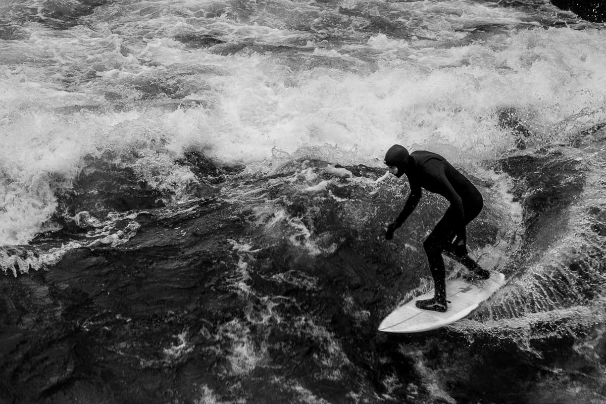 Surfer auf seinem Board in Ganzkörperneopren mit Kaputze auf der Eisbachwelle im Englischen Garten in München - Dezember 2022.