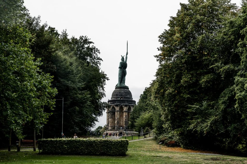 Sichtachse zum Hermannsdenkmal vom Bismarckstein aus - mittig das Hermannsdenkmal, rechts und links hohe, große Bäume, eine Hecke im vorderen Bereich.