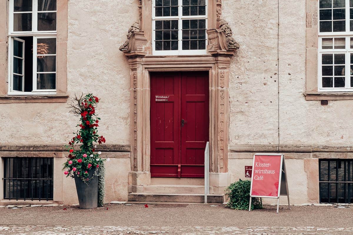 Eingang zum Klosterwirtshaus und Café sowie Brauerei des Kloster Dalheims mit entsprechender Beschriftung der Tür und Aufsteller rechts neben der Tür. 
Blumenarrangement links neben der Tür. 
