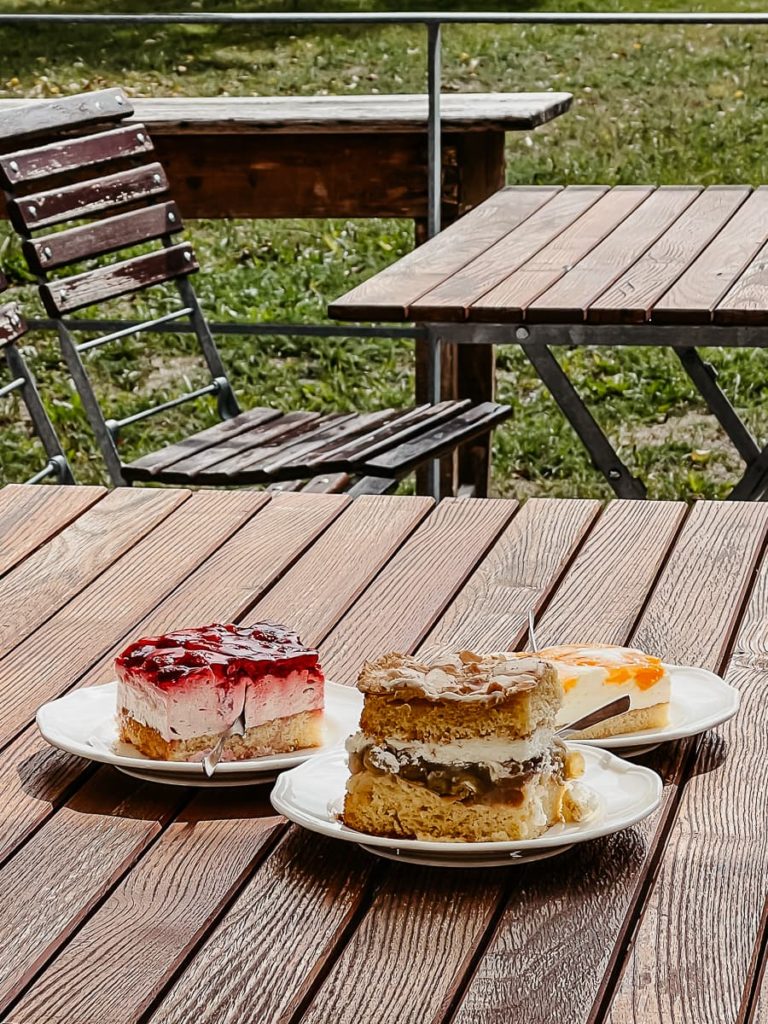 Drei verschiedene Kuchenstückchen aus der Klostergastronomie auf einem Lattenklapptisch auf der Außenterrasse im Kloster Dalheim, dahinter leerer Klappstuhl und weiterer Tisch mit Holzlatten. 