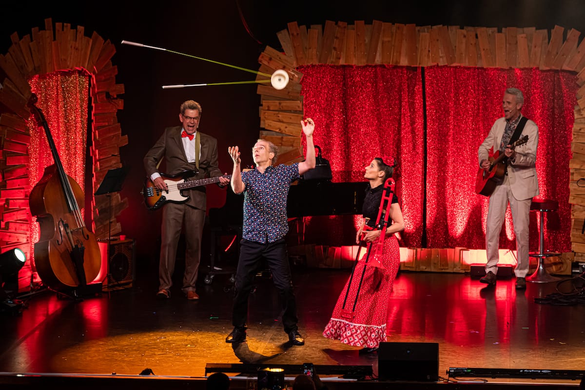 Zwei Musiker singen im Hintergrund, beide im Anzug, links mit Fliege, rechts mit Einstecktuch. Fer links spielt E-Bass, der rechts Akkustikgitarre. Davor die Akrobaten des Duos E1nz - Jonas links wirft gerade sein Diabolo mit Seil hoch, rechts daneben steht Ester im Kleid mit einer Posaune und sieht Richtung Diabolo. 