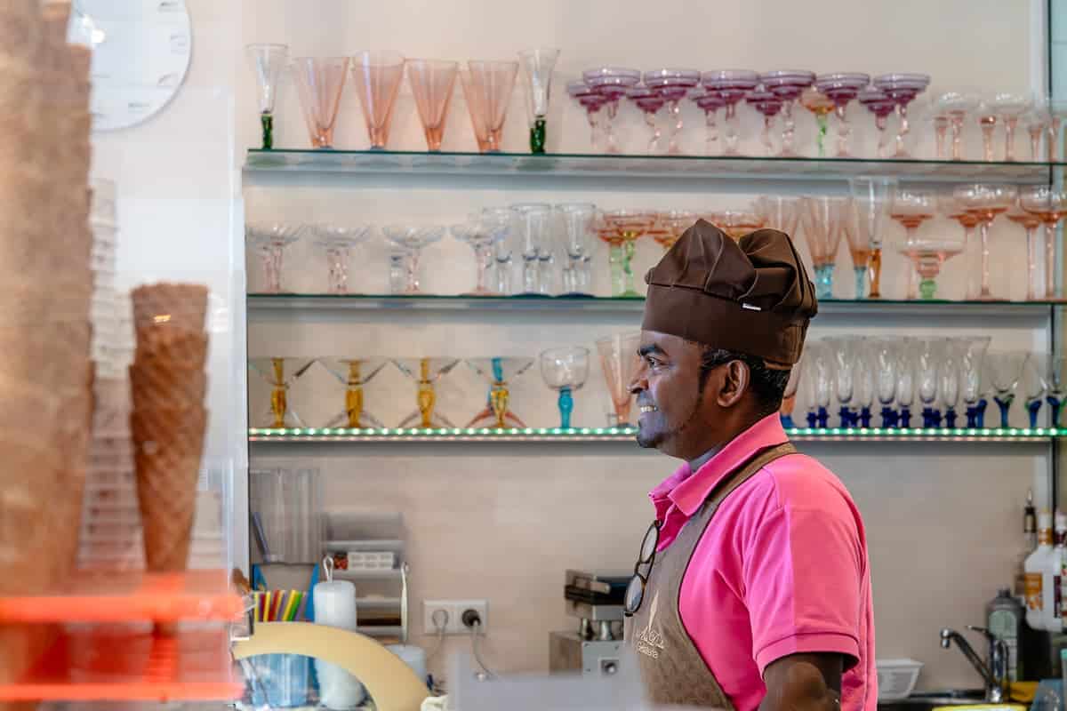 Thavakumar Maniam hinter der Theke in seinem Eisladen Arte Dolce Gelateria, Karlsruhe. Er schaut lächelnd hinter der Theke hervor mit Schürze und Mütze, im Regal dahinter verschiedene Gläser für Eiskreationen, links vorne gestapelte Eiswaffeln.
