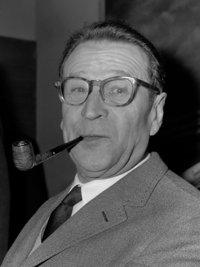 Portrait von Georges Simenon aus dem Jahr 1965, linke Schulter vorne, Pfeife im rechten Mundwinkel, Hornbrille. Anzug mit Krawatte. Foto via Wikimedia Commons & Nationaal Archief, CC0 1.0-Lizenz