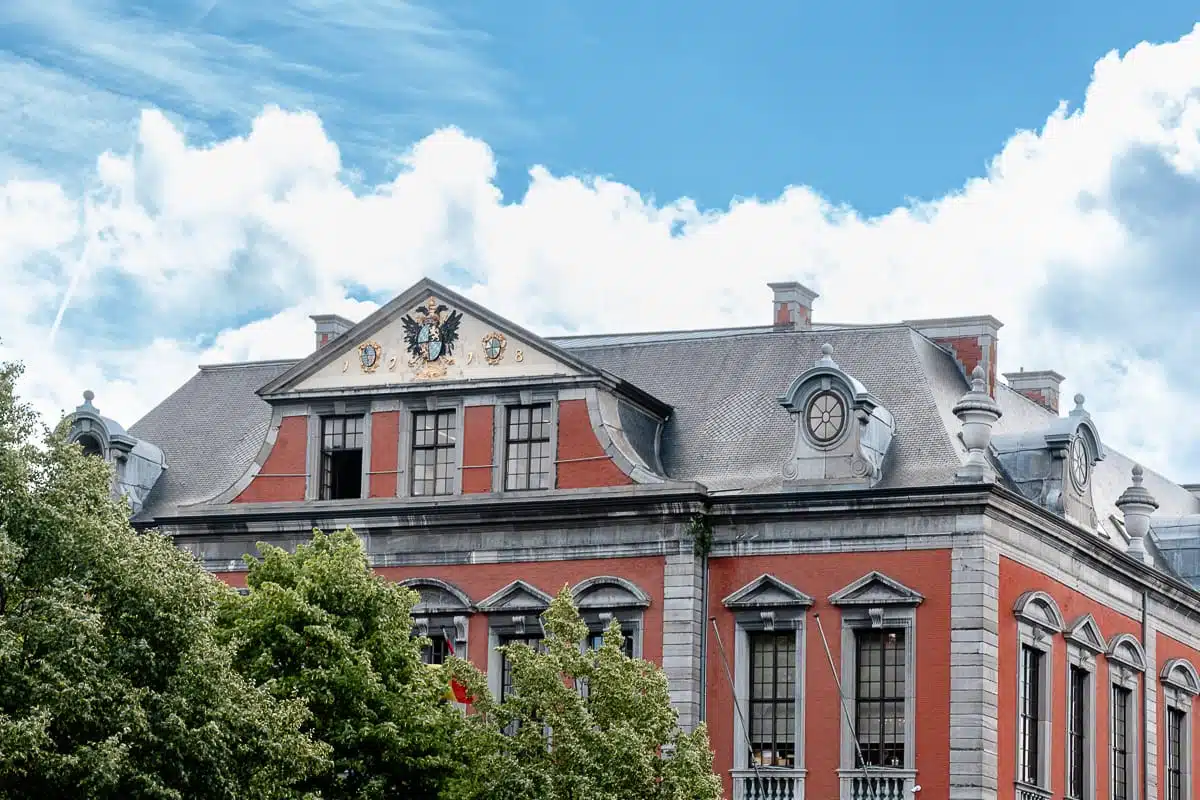 Ein historisches Gebäude aus rotem Backstein mit hohen Fenstern, kunstvollen Steinverzierungen und einem dreieckigen Giebel mit einem schwarzen Doppeladler als Wappen - das Rathaus von Lüttich, das La Violette. Grüne, belaubte Bäume verdecken teilweise den unteren Teil.