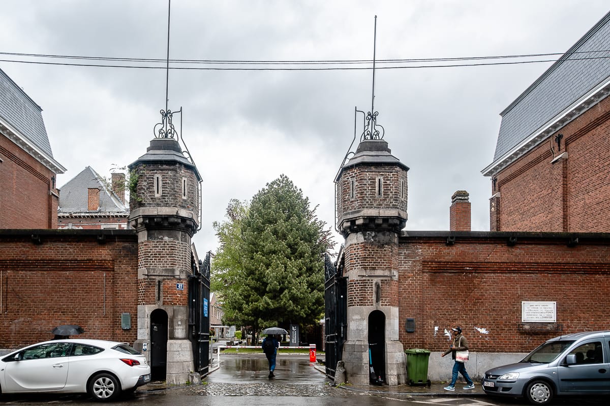 Eingang zur Caserne des Lanciers Fonck. Eingang umrahmt von zwei Wachtürmchen mit Einlässen für die Soldaten, die den Eingang bewachten. heute offen, rechts und links parken Autos, eine Person betritt gerade das Gelände.