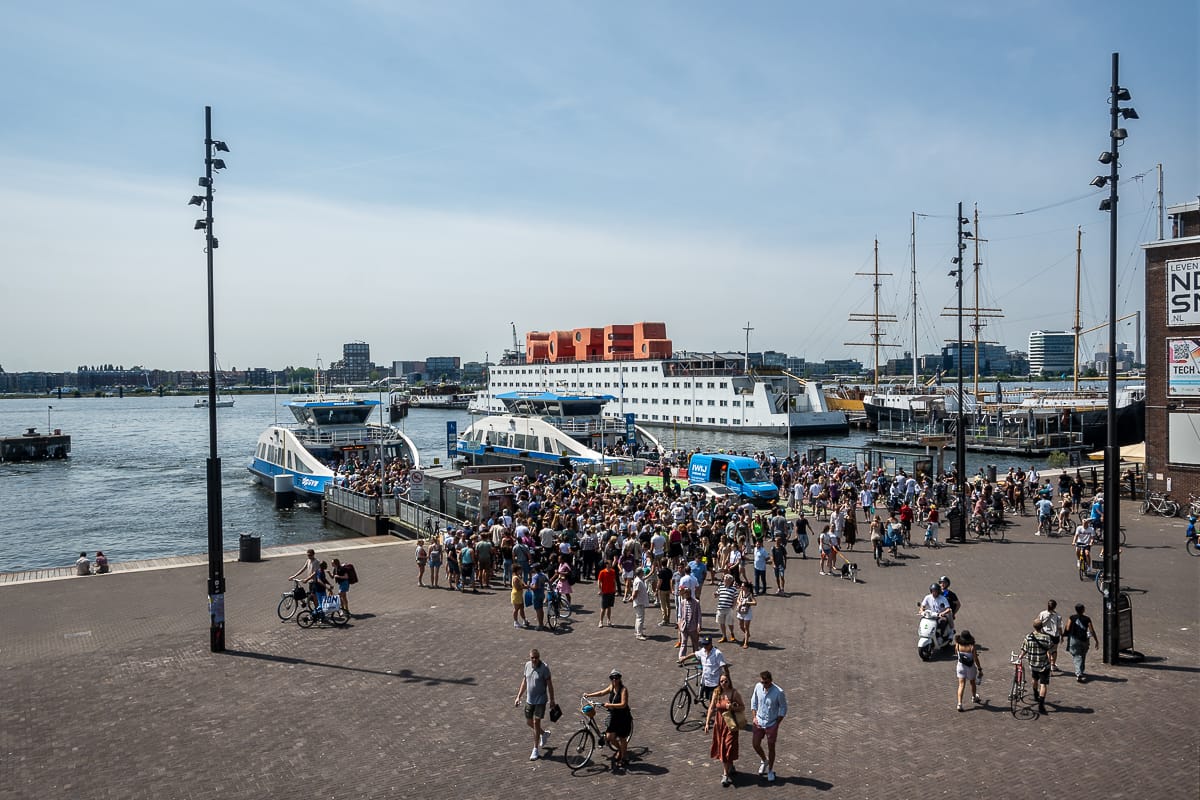 Menschen strömen aus zwei Fähren an der Anlegestelle NDSM Werf in Amsterdam-Noord, dahinter das botel.