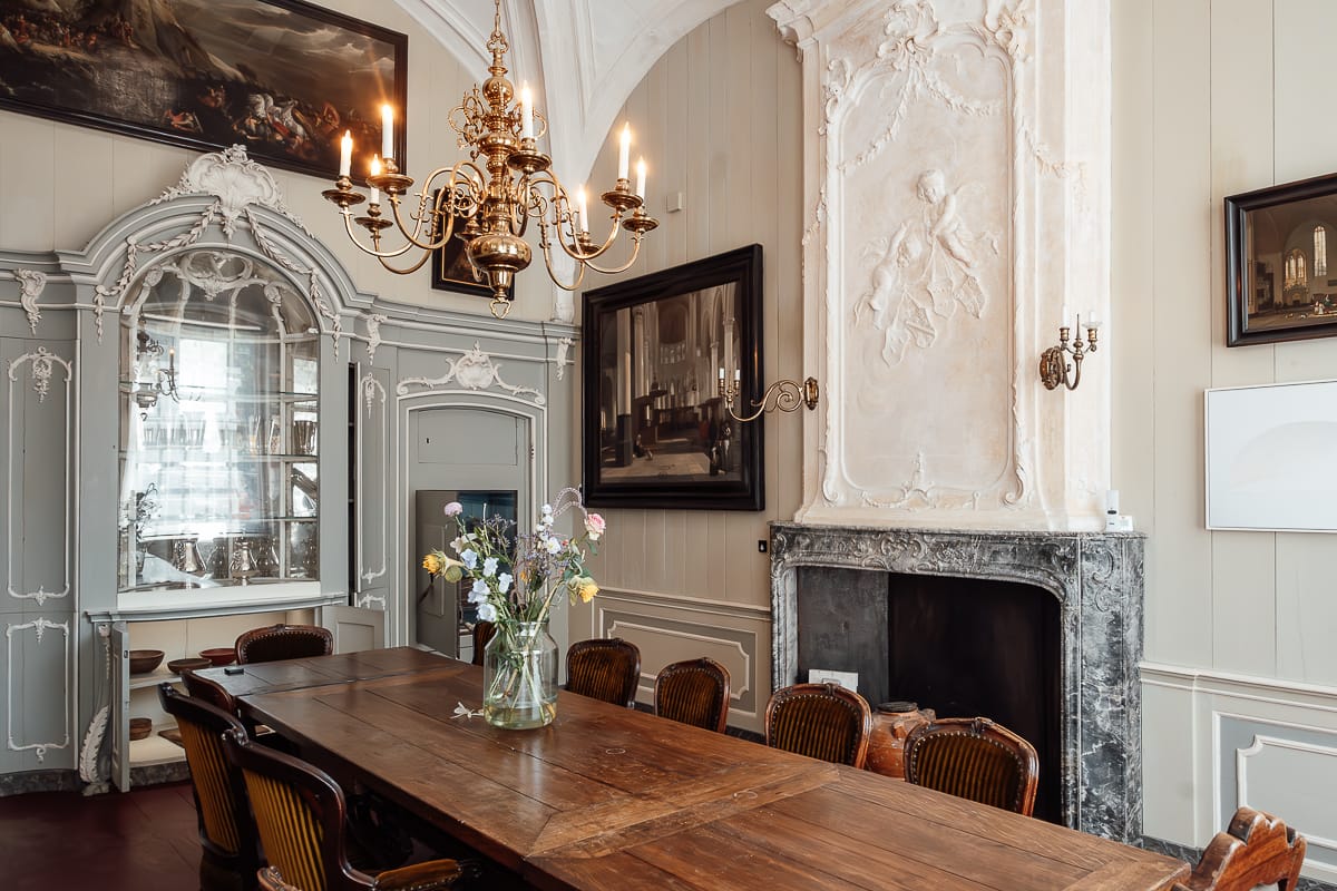 Büro des Kirchenvorstehers mit Holztafel und mehreren Stühlen, Blumenvase darauf, Kamin dahinter, verzierter Stuck an der Wand, großen Bild, Kronleuchter über der Tafel.