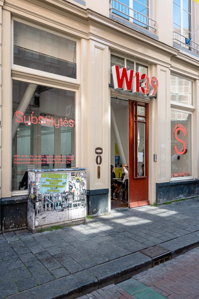 Außenansicht des W139 Amsterdam, Schriftzug über der Tür, deren linker Flügel ist geöffnet, links und rechts Schaufenster mit Schriftzügen zur aktuellen Ausstsellung.