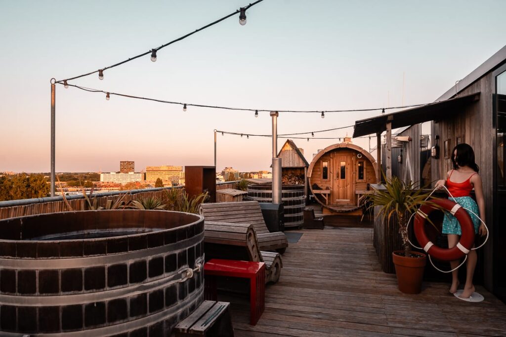 Sauna (ein großes Holzfass mit Fenstern und Tür) und Hot Tubs im obersten Stock, Volkshotel Amsterdam. Holzboden, Sonnenuntergang, Lichterketten quer gespannt.