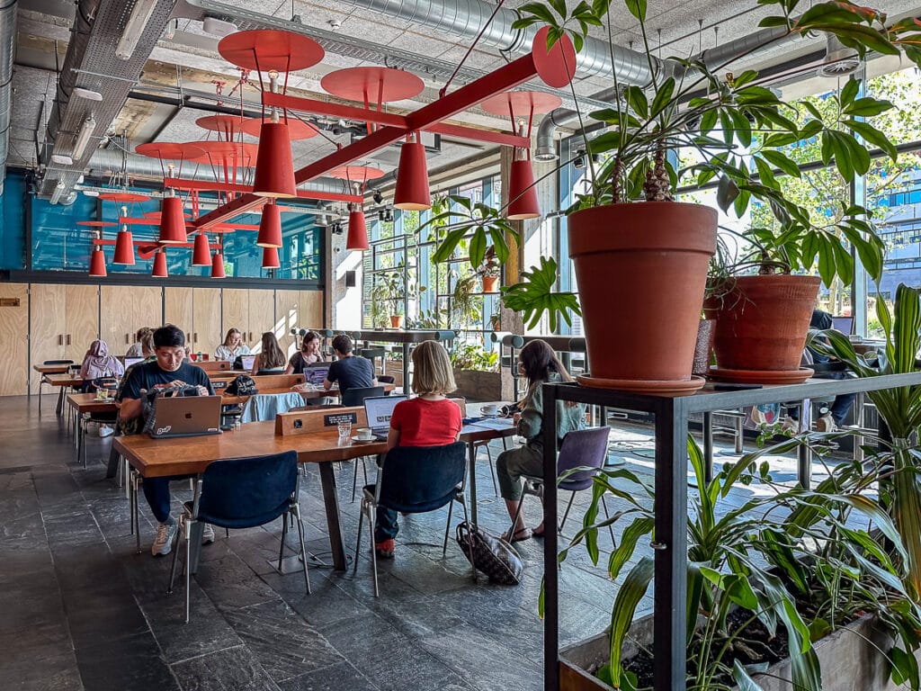 Coworking Bereich im Volkshotel Amsterdam - Tische an denen Menschen mit Laptop sitzen und arbeiten, Lampen darüber, im Vordergrund rechts Regal mit Pflanzen.