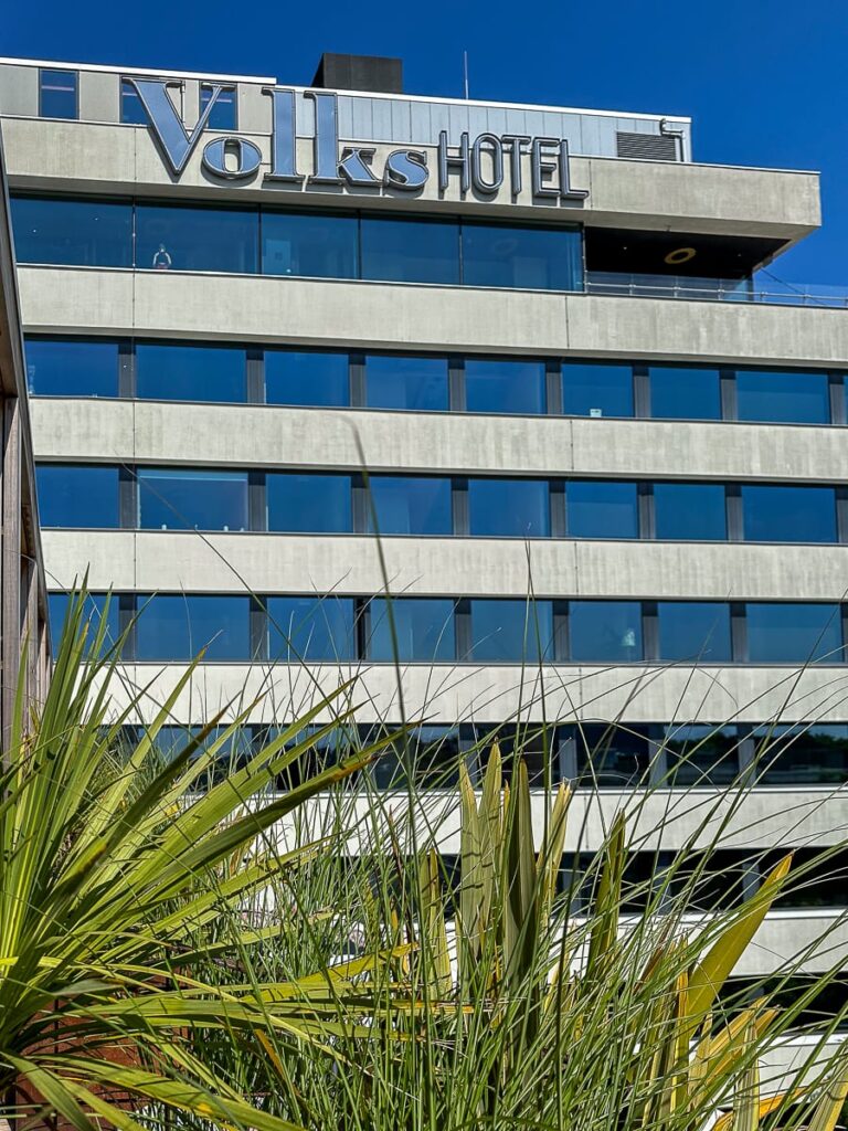 Ansicht des Volkshotels - Beton und Fensterfronten wechseln sich ab, eine grüne Palmenpflanze im Vordergrund, oben am Gebäude Schriftzug "Volkshotel" in zwei verschiedenen Schriftarten.