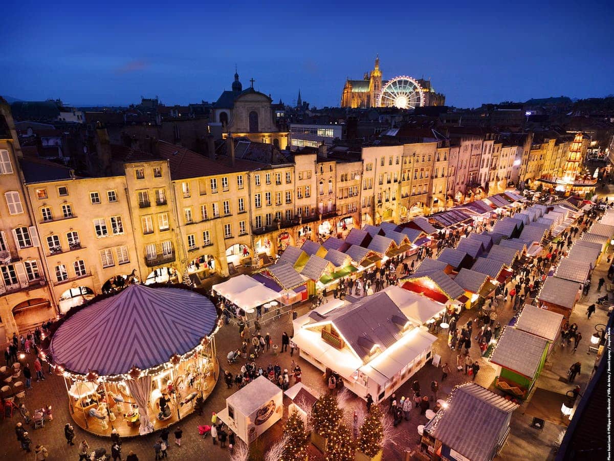 Marché de Noël Place St Louis - Ansicht von oben auf den Markt zur blauen Stunde mit beleuchteten Ständen und Karusssell, Kathedrale mit Riesenrad im Hintergrund.
Foto © Ph.Gisselbrecht / Ville de Metz / Benoit Lapray / Inspire Metz