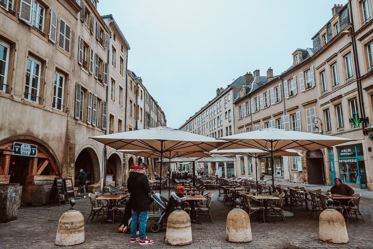 Am Place Saint-Louis, zwischen mittelalterlichen Häusern mit Arkaden, Außengastromöbel und Schirme sind aufgestellt, 2-3 Menschen sitzen und stehen davor.