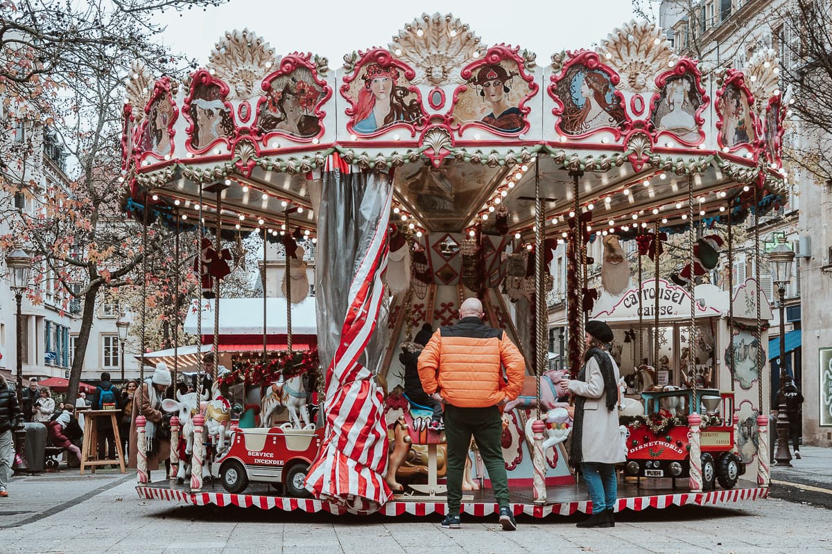 Karussell auf dem Place Saint-Jacques, Weihnachtsmarkt Metz. Eltern stehen vor dem Karussell und ihr Kind sitzt auf einem der Tiere des Karussels. 