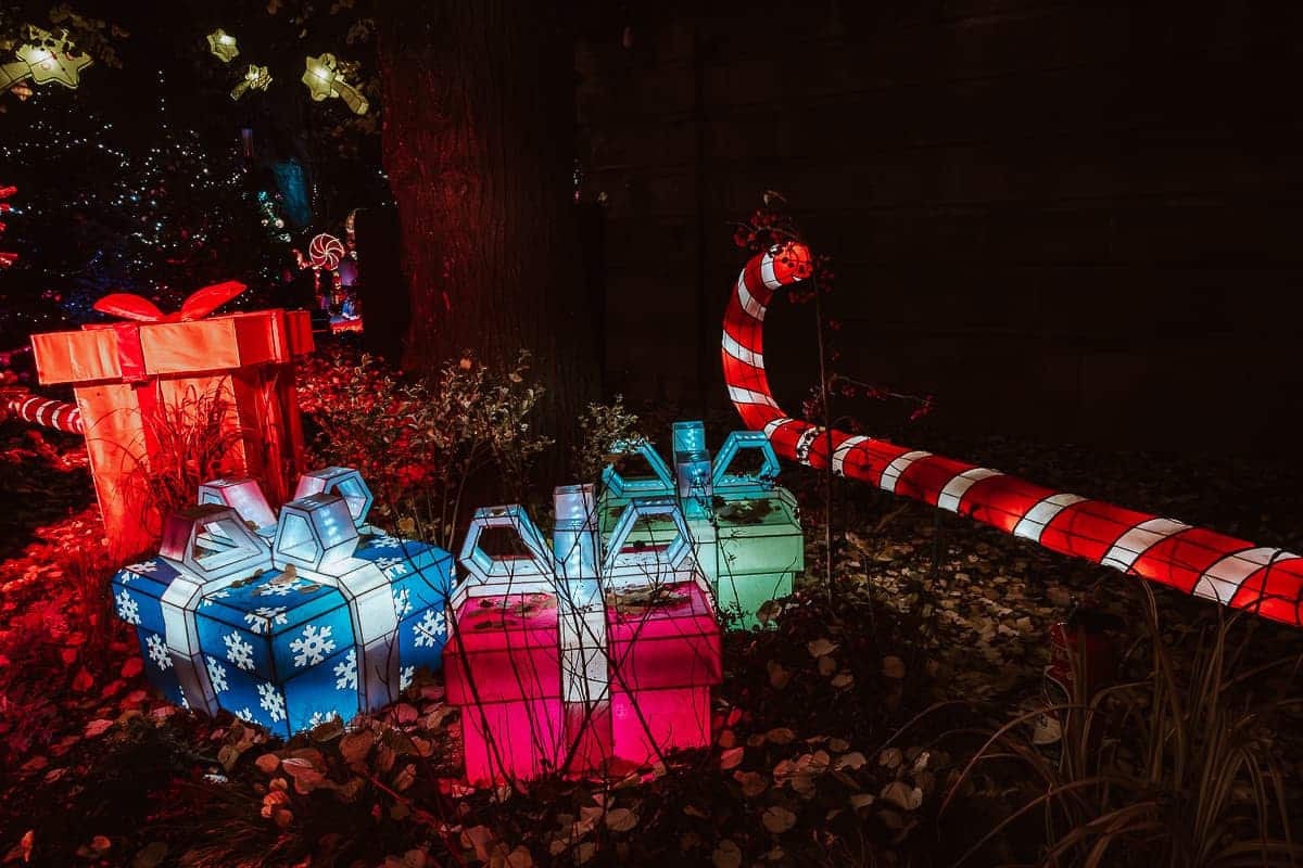 Inmitten der bezaubernden Nachtszene, die an einen Weihnachtsmarkt in Metz erinnert, ziehen große leuchtende Geschenkboxen in Rot, Blau und Grün, die mit Schneeflockenmustern verziert sind, die Zuschauer in ihren Bann. Ein riesiger beleuchteter Zuckerstangenbogen wölbt sich durch den Hintergrund und wirft einen warmen Schein zwischen gefallenen Blättern und Bäumen.