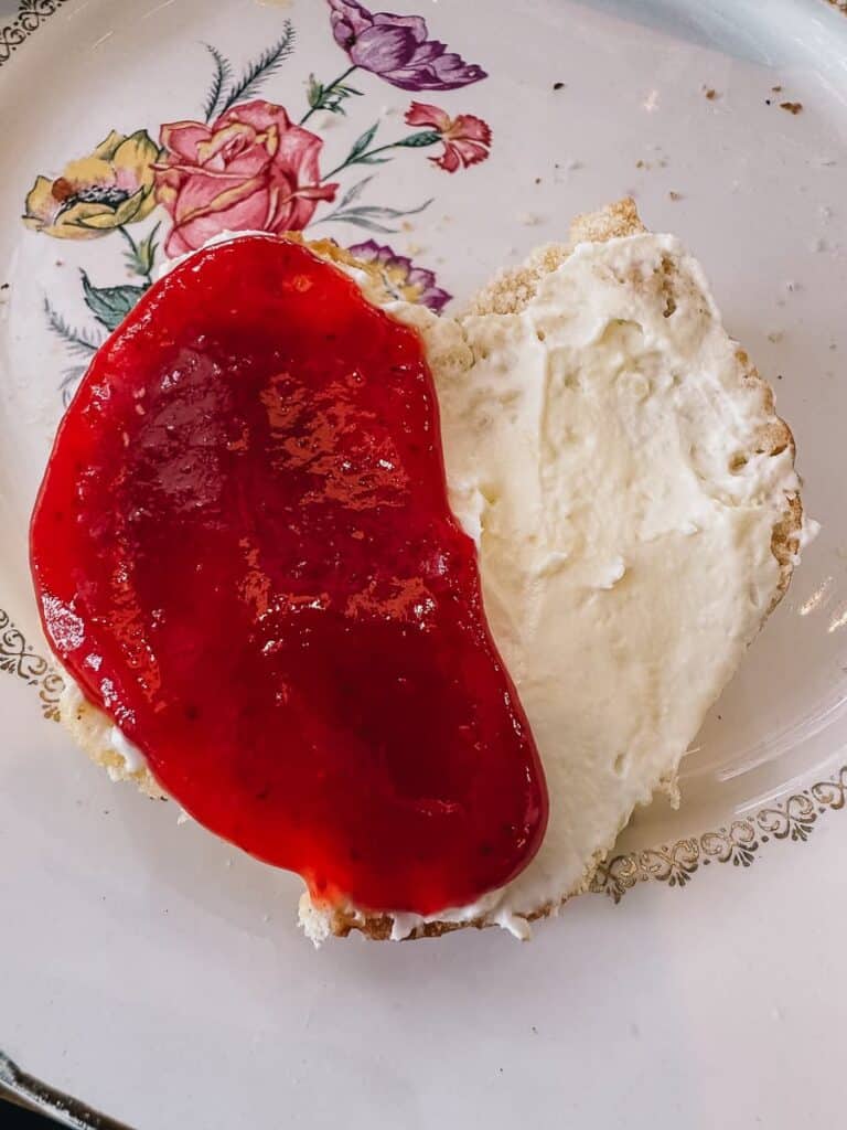 Ein Scone bestrichen links mit Marmelade, rechts mit clotted cream auf einem bemalten Teller (Blumenmotiv).