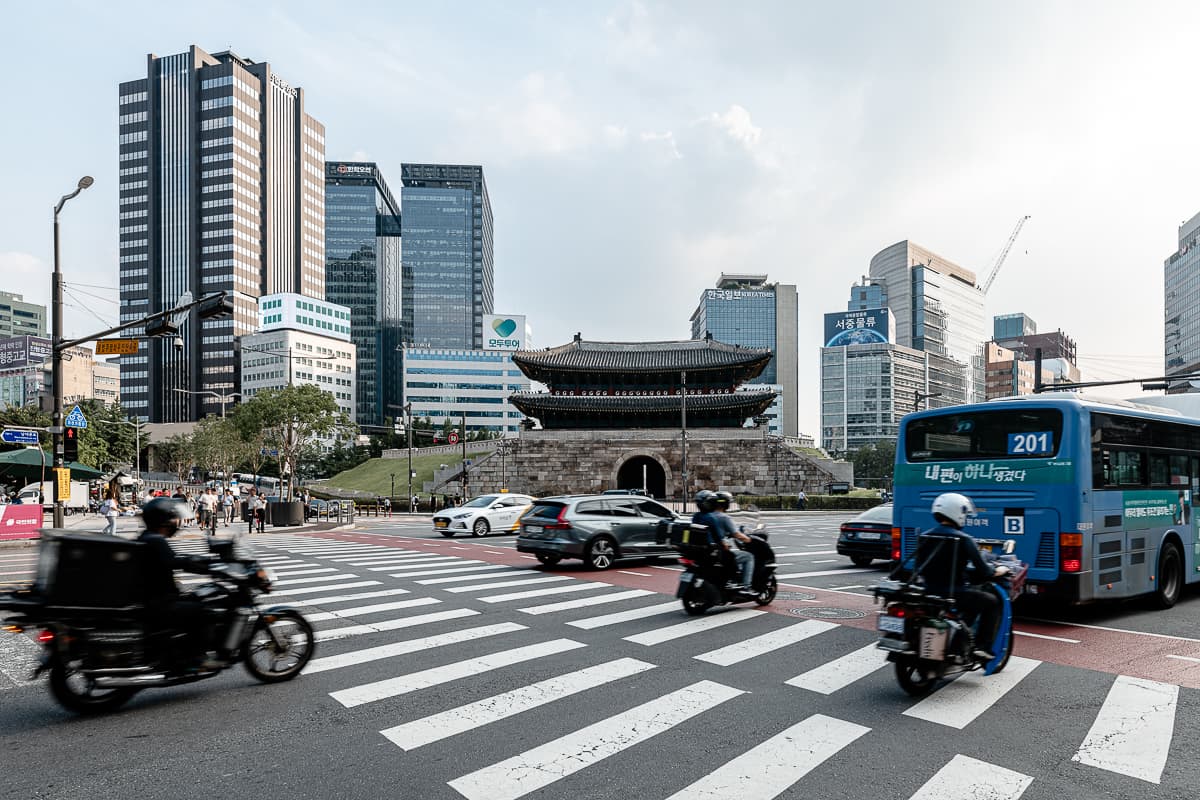 Sungnyemun Tor in Seoul mit Hochhäusern dahinter und großer Kreuzung davor. Diese Kreuzung wird von Motorrädern und einem Bus von links nach rechts überquert. 