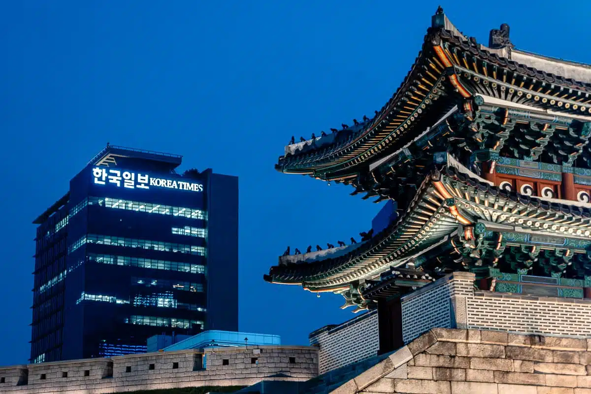 Ausschnitt des Sungnyemun-Tors in Seoul in der Dämmerung, im Hintergrund ein modernes Hochhaus mit Schriftzug "Korea Times".