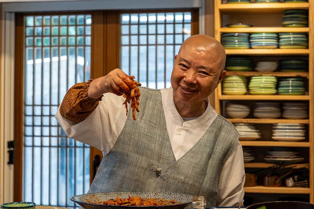 Die kochende Nonnen Jeong Kwan in ihrer Küche im Baegyangsa-Kloster im Jeollanam-do, die in die Kamera lächelt, die rechte Hand mit Essen über der enstprechenden großen Schüssel. 