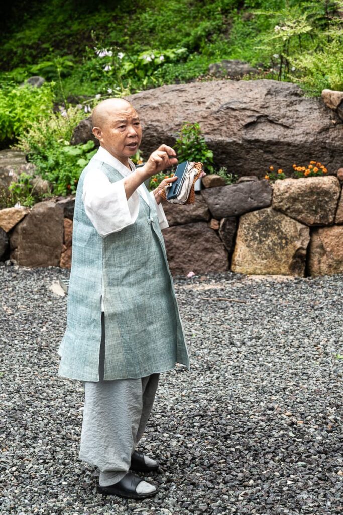 Buddhistische Nonne Jeong Kwan in lockerer Tempelkleidung - Hose, Bluse, längere Weste, dazu Socken in einfachen Sandalen, in der linken Hand Smartphone und Kochärmel zum Überziehen, steht auf einem Schotteruntergrund, dahinter Garten der Eremitage in Jeollanam-do zu sehen. Gestikuliert mit rechts und ruft einer anderen Person etwas zu.