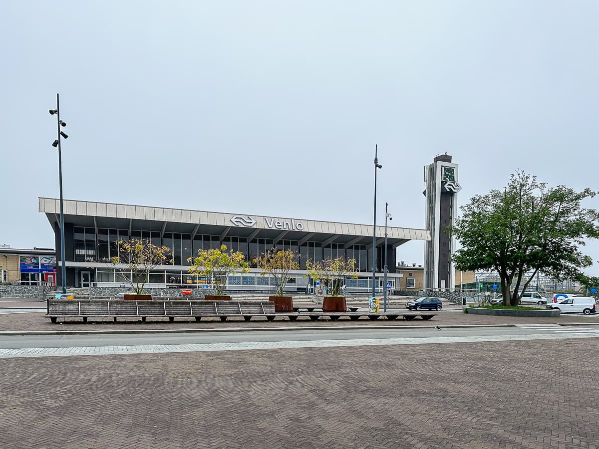 Blick vom Bahnhofsvorplatz auf den Bahnhof Venlo, ein schmuckloser Zweckbau mit einer Art Uhrturm daneben. Auf dem Vorplatz einige kleine Bäume und eine Reihe an Sitzbänken. 