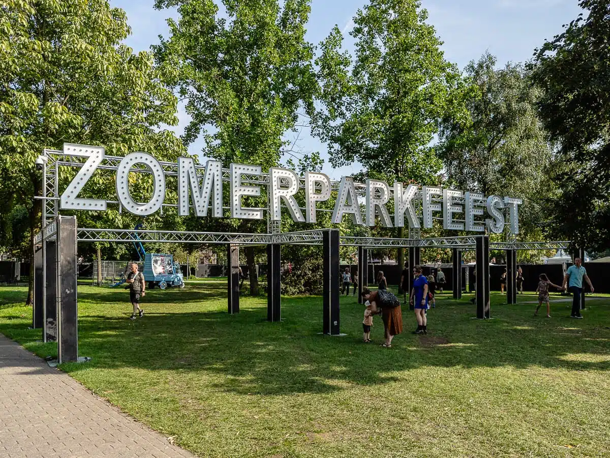 Eingang zum Zomerparkfeest mit riesigen, beleuchteten Buchstaben auf Stahlträgern, darunter Eltern und Kinder, Gäste, die zum und vom Gelände strömen.