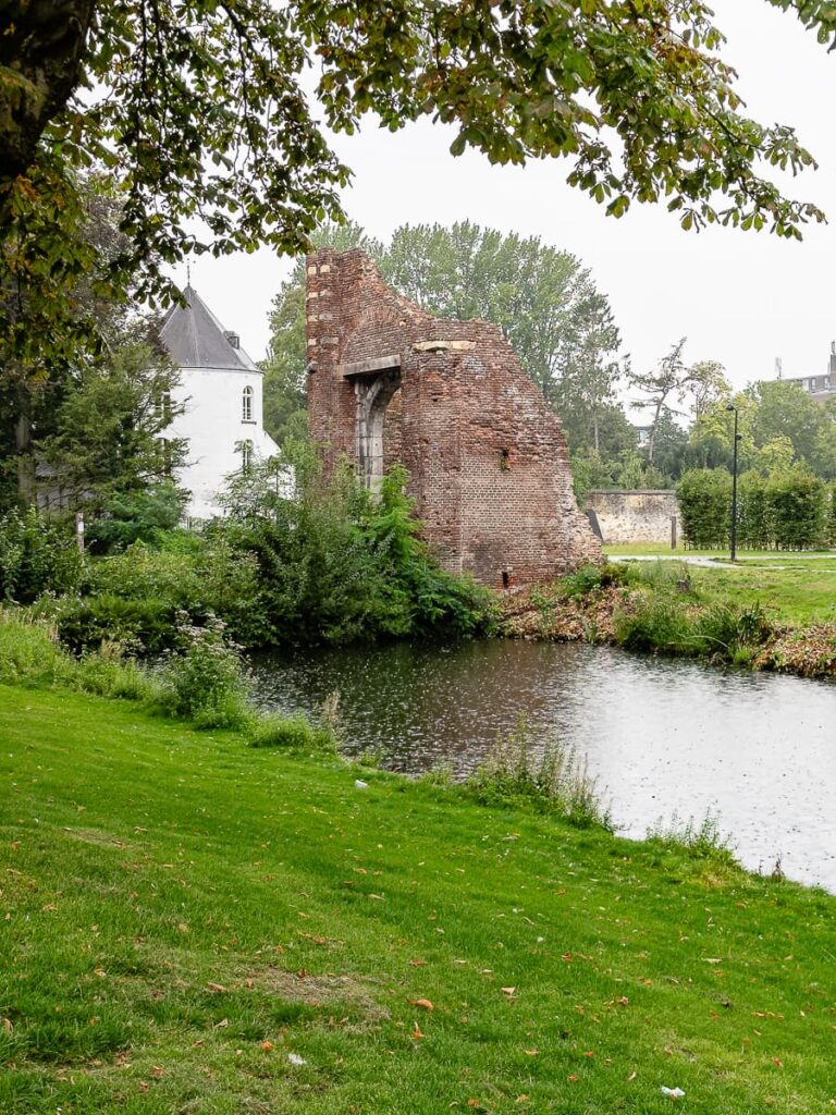 Überreste, neueres Schlösschen dahinter und künstlichem Wassergraben davor, gerahmt von Baumzweigen.