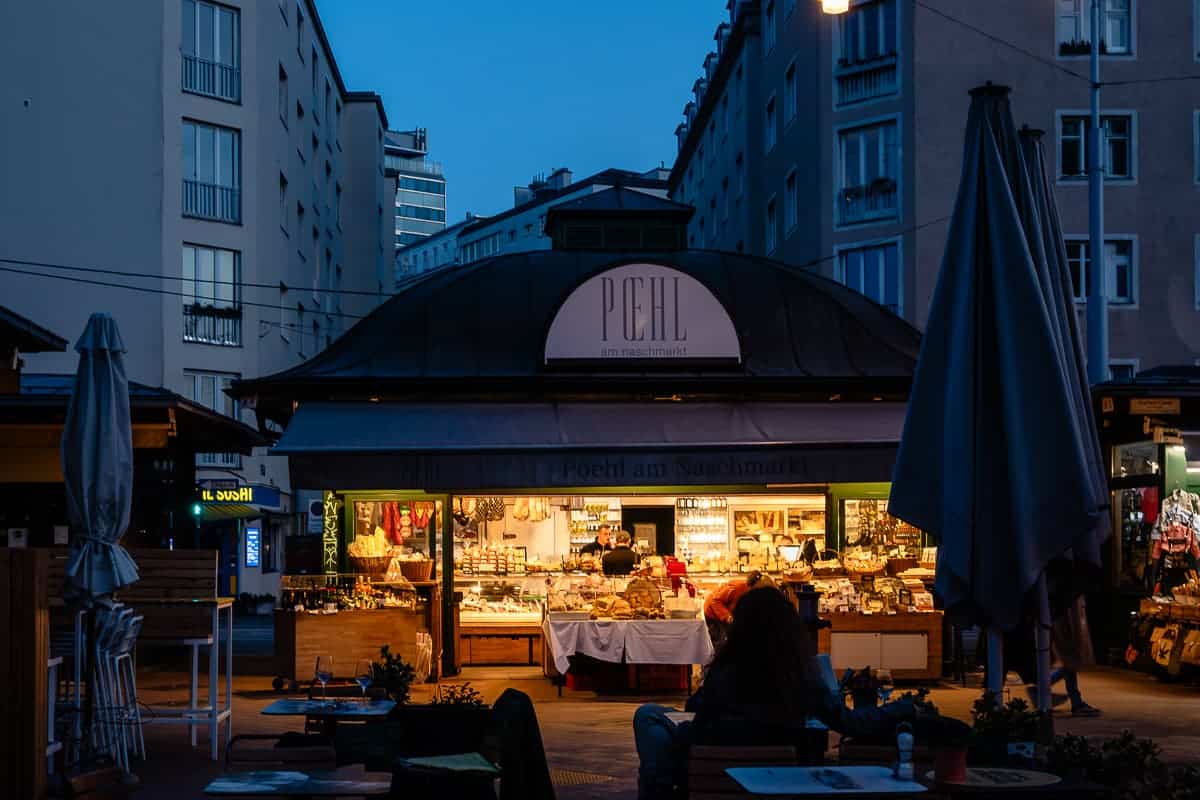 Während der blauen Stunde, in den Abendstunden auf dem Naschmarkt in Wien. Der beleuchtete Stand, ein kleines Häuschen von "Poehl am Naschmarkt", davor einige Tische, zusammengefaltete Schirme, eine Person sitzt an einem Tisch und genießt ihren Wein.