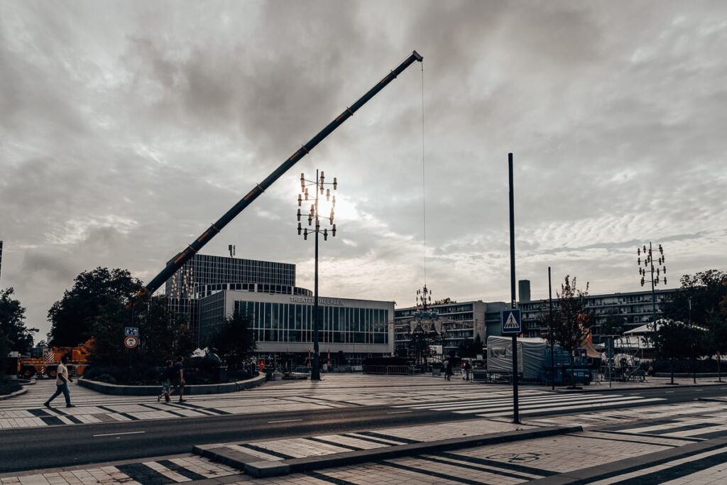Kranaufbau vor dem Theater Heerlen, einem großen Platz. Kran steht links und hält ein aus Metall gefertigtes Dach , das über einem offenen Pavillon rechts schwebt. Dahinter links das Theater, rechts ein Spiegelzelt und andere Zelte/Aufbauten für das Festivals, Getränkestände, Tontechnik sowie Absperrungen. 