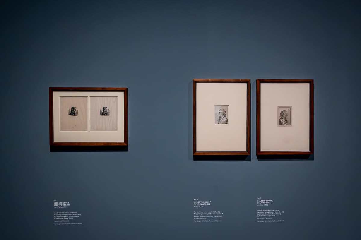 Selbstbildnisse von Caspar David Friedrich, Kunsthalle Hamburg. Drei gerahmte Bilder, links Querformat, rechts zwei Hochformat. Hochformate schauen in entgegengesetzte Richtungen, im Querformat sind zwei Bildnisse, beide schauen nach rechts. Darunter Beschreibungstexte zu den Bildern.