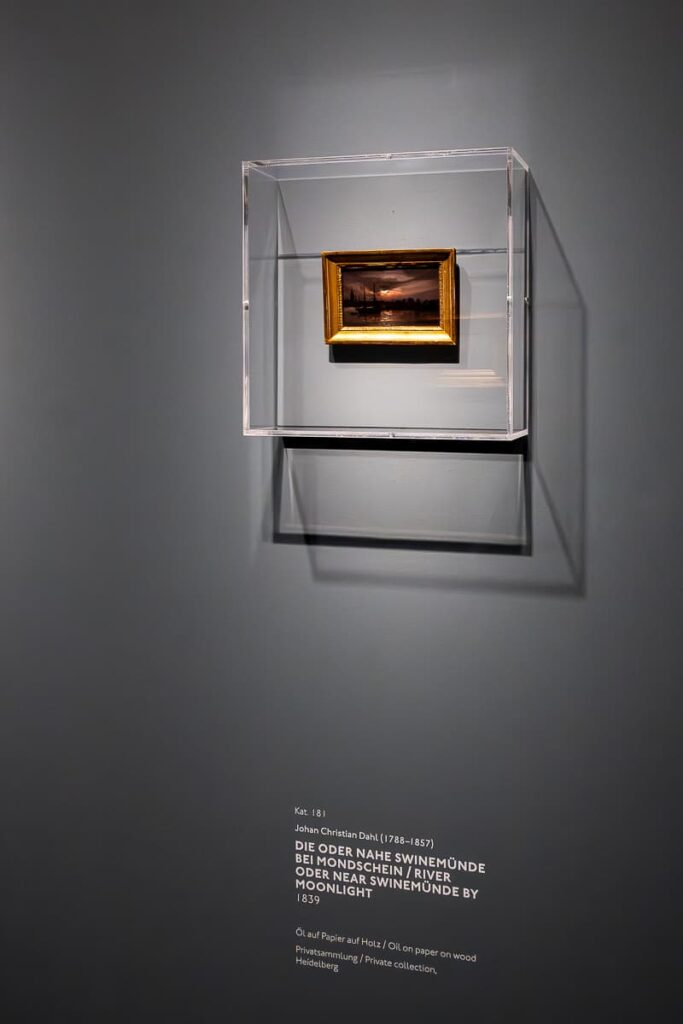 Hochformatiges Foto mit einem sehr kleinen Bild in einem dezenten Goldrahmen hinter Plexiglas, darunter Begleittext zum Bild. Johan Christian Dahl, Die Oder nahe Swinemünde bei Mondschein, 1839, Öl auf Papier auf Holz, Privatsammlung Heidelberg in der Ausstellung Caspar David Friedrich, Kunsthalle Hamburg.