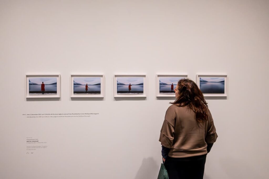 Eine Frau steht rechts vor eine Reihe von fünf Fotos und betrachtet die linken Fotos, kleinformatig, weiße Rahmen. Swaantje Güntzel, Arctic Yoghurt, 2. Dezember 2021, Intervention Ofotfjord, Norwegen, 5 Fogorafien, Courtesy of the artist.