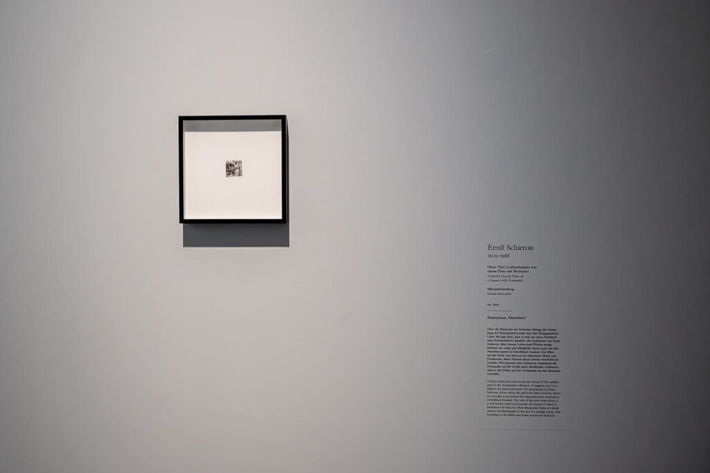 Sehr kleines Bild in einem quadratischen Rahmen an einer Wand. Beschreibungstext und Details sehr weit rechts davon. Bild: Ernst Schieron, "Ohne Titel (Luftaufnahme von einem Platz mit Brunnen)", ca. 1935, Silbergelatineabzug (Fragment einer Postkarte), 3,2x3,4 cm, Kunstpalast Düsseldorf.