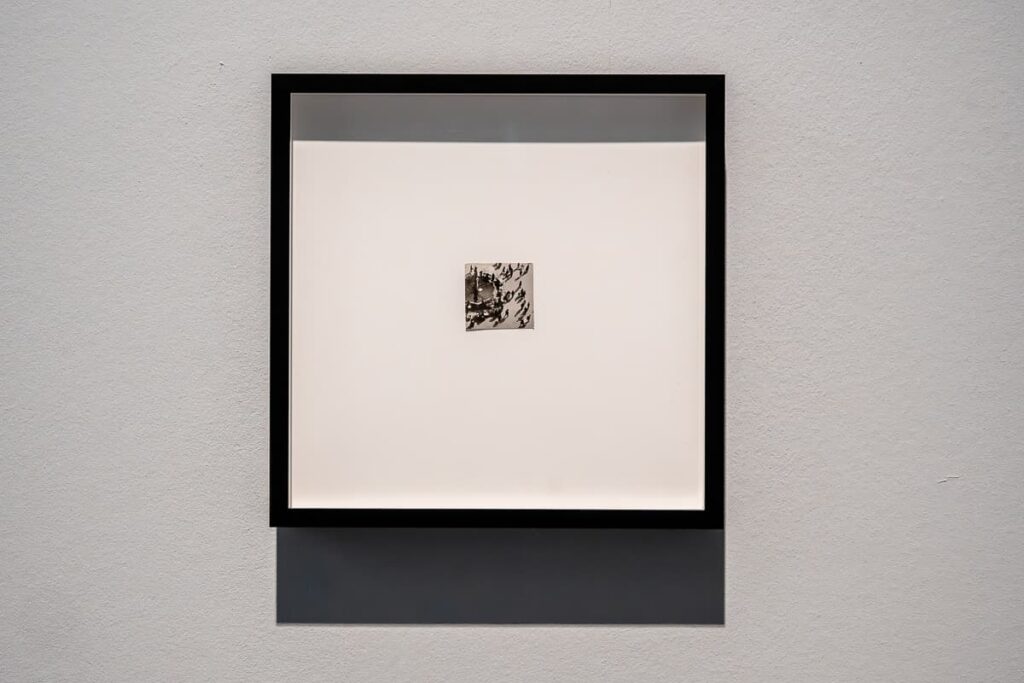 Sehr kleines Bild in einem quadratischen Rahmen an einer Wand. Nahaufnahme. Bild: Ernst Schieron, "Ohne Titel (Luftaufnahme von einem Platz mit Brunnen)", ca. 1935, Silbergelatineabzug (Fragment einer Postkarte), 3,2x3,4 cm, Kunstpalast Düsseldorf.