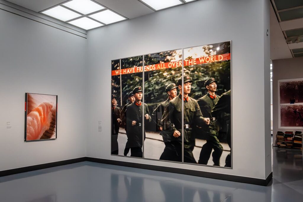 Blick in eine Ecke der Ausstellung, sehr großes Foto, Querformat von Katharina Sieverding an einer Zwischenwand, rechts daneben Durchgang zum nächsten Raum. Bild zeigt joggende, chinesische Soldaten, die von links nach rechts laufen, darüber ein Spruchband "We have friends all over the world". An der linken Wand ein kleineres, hochformatigeres Bild mit Sushi? Leider nicht im Katalog vermerkt.