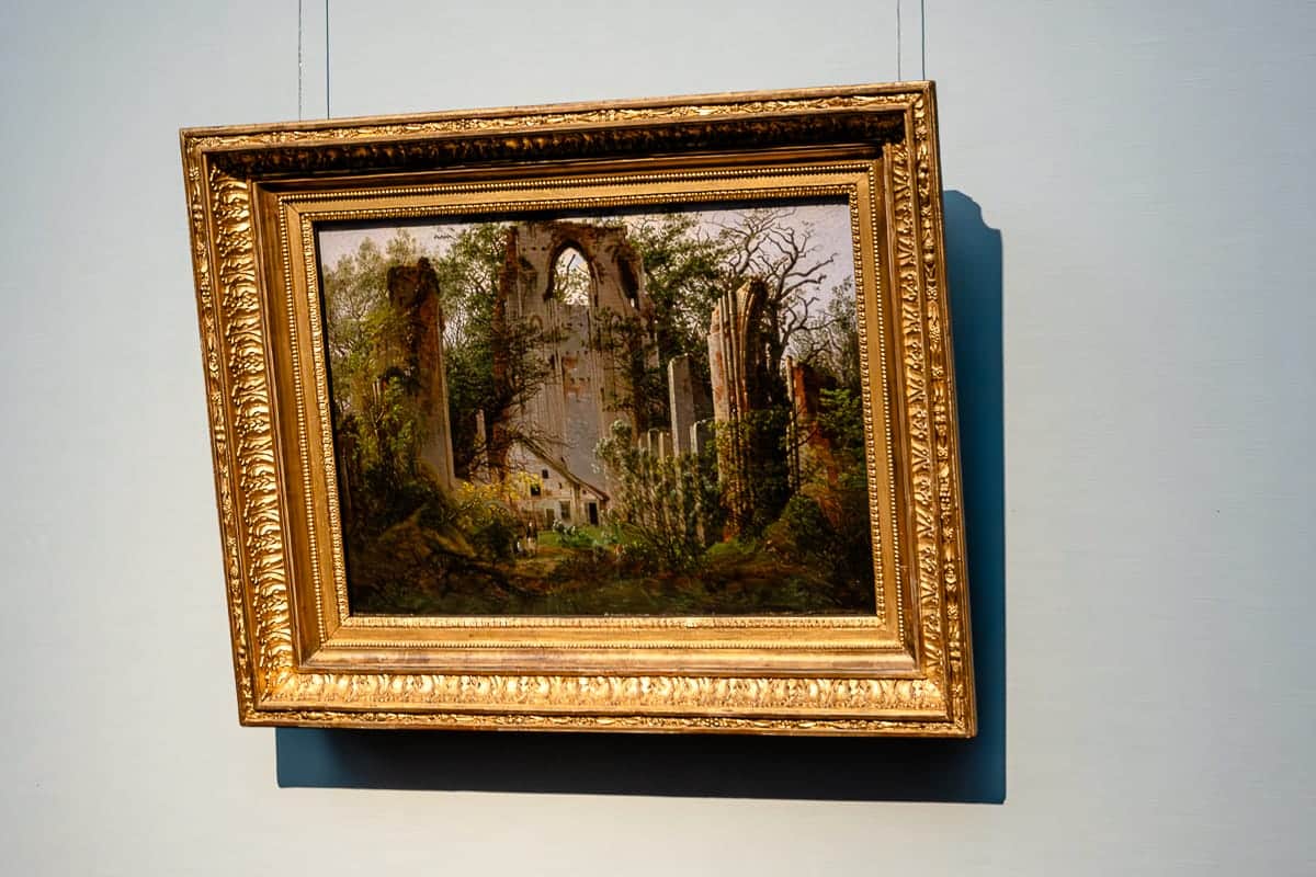 Querformatiges Bild "Klosterruine Eldena bei Greifswald" (1824/25) von Caspar David Friedrich, Klosterruine ziemlich überwuchert mit vielen Bäumen und Gründ, dazwischen/darunter ein verlassenes Haus.