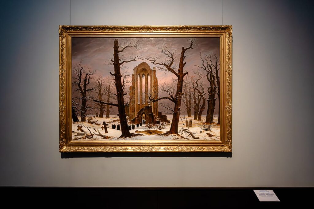 Eine Klosterruine im Schnee, eingerahmt von zwei großen Bäumen, karg, ohne Blätter, weitere Bäume im Hintergrund. Alte Grabsteine unregelmäßig verteilt, Menschen gehen auf die Ruine zu und durchschreiten einen Eingang. Farbig. Kopie eines unbekannten Malers nach 1855 von "Klosterfriedhof im Schnee" von Caspar David Friedrich.